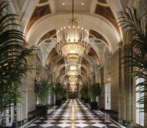 A hallway at the Waldorf Astoria hotel with silver chandeliers and a black and white tiled floor.