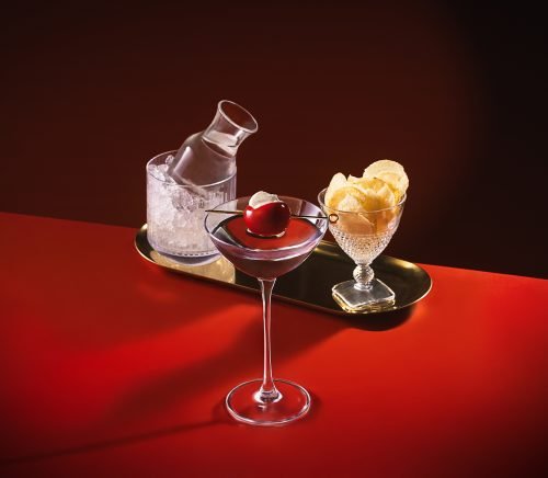 A cocktail next to a glass full of potato chips and a pitcher of water set to a red background