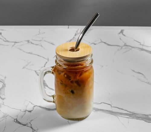 A latte in a glass cup with a handle, lid and straw.