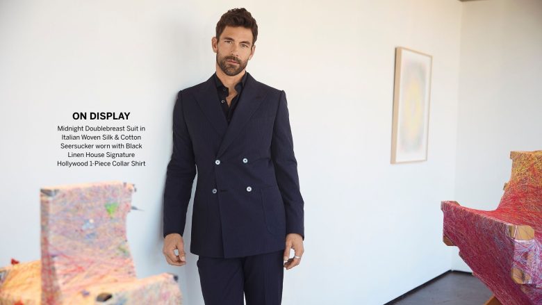 A man leans against a wall in an art gallery wearing a double breasted suit.