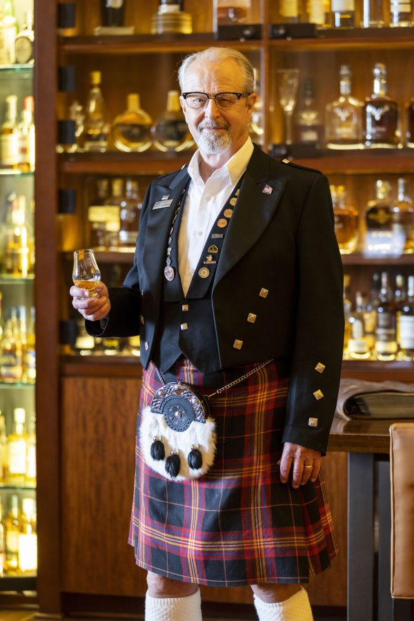 A man poses in a kilt holding a glass of Scotch.